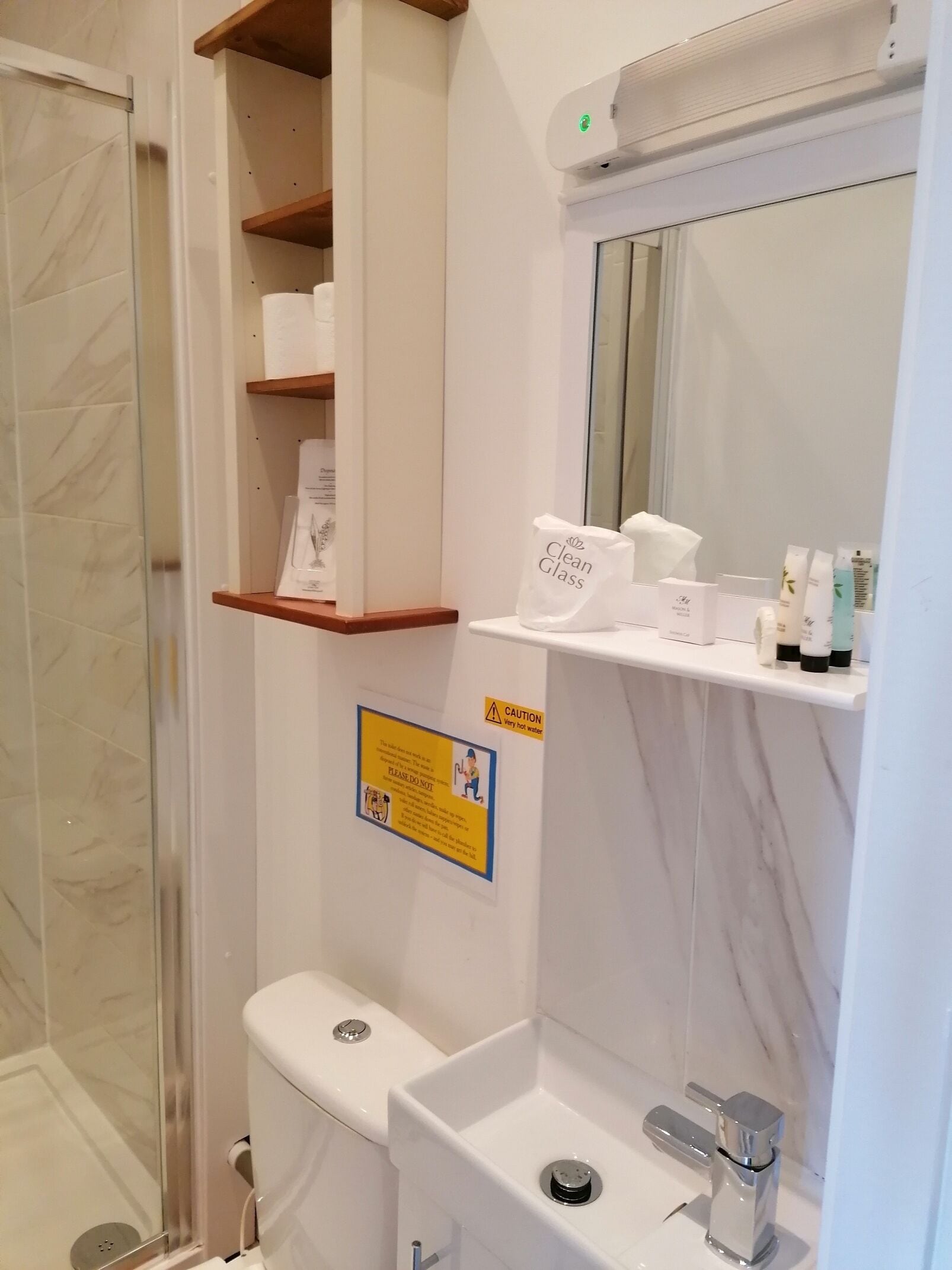 Baño con inodoro, lavabo y zona de ducha. Se observan repisas con artículos de tocador y un letrero de precaución.