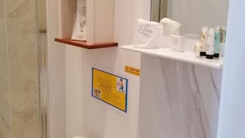 Baño con inodoro, lavabo y zona de ducha. Se observan repisas con artículos de tocador y un letrero de precaución.