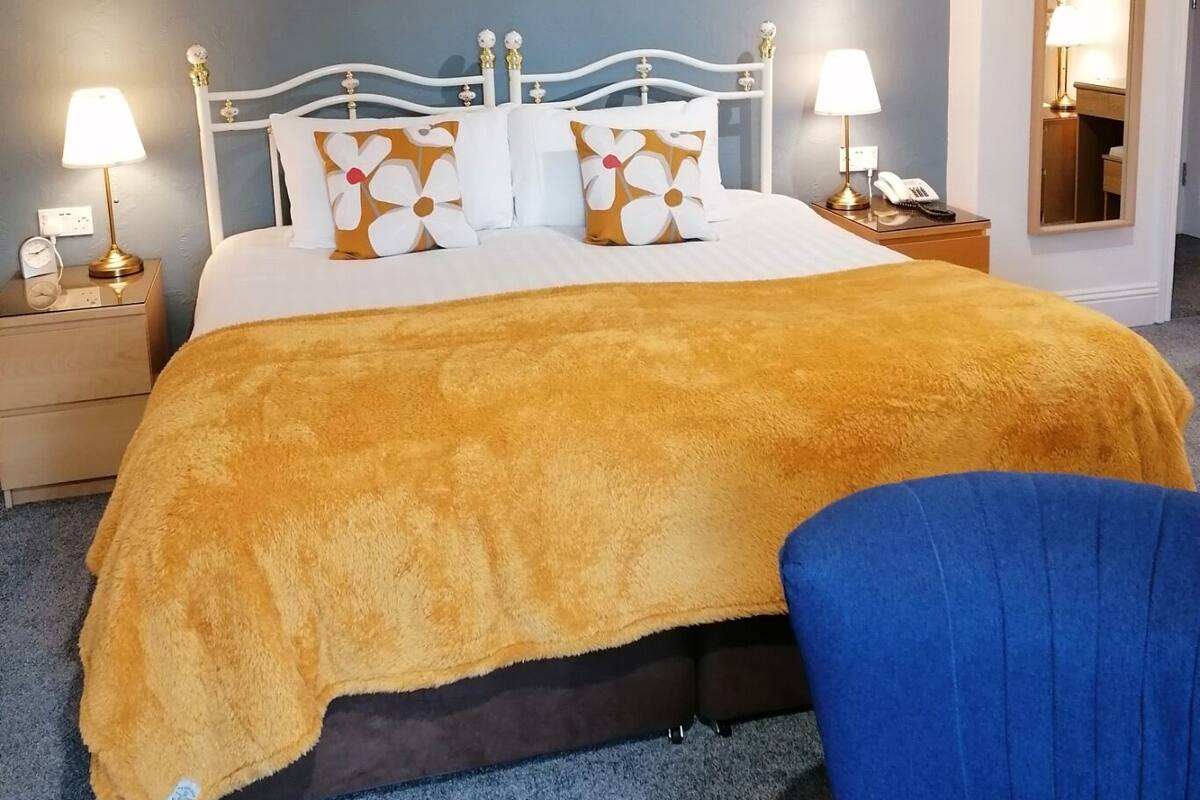 Um quarto com uma cama, duas mesas de cabeceira, um criado-mudo, uma cadeira azul e um espelho na parede.