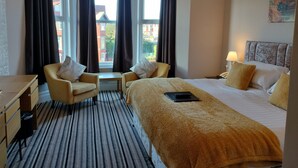 Habitación de hotel con una cama, dos sillones, una mesita, una lámpara y vistas a edificios a través de amplias ventanas.