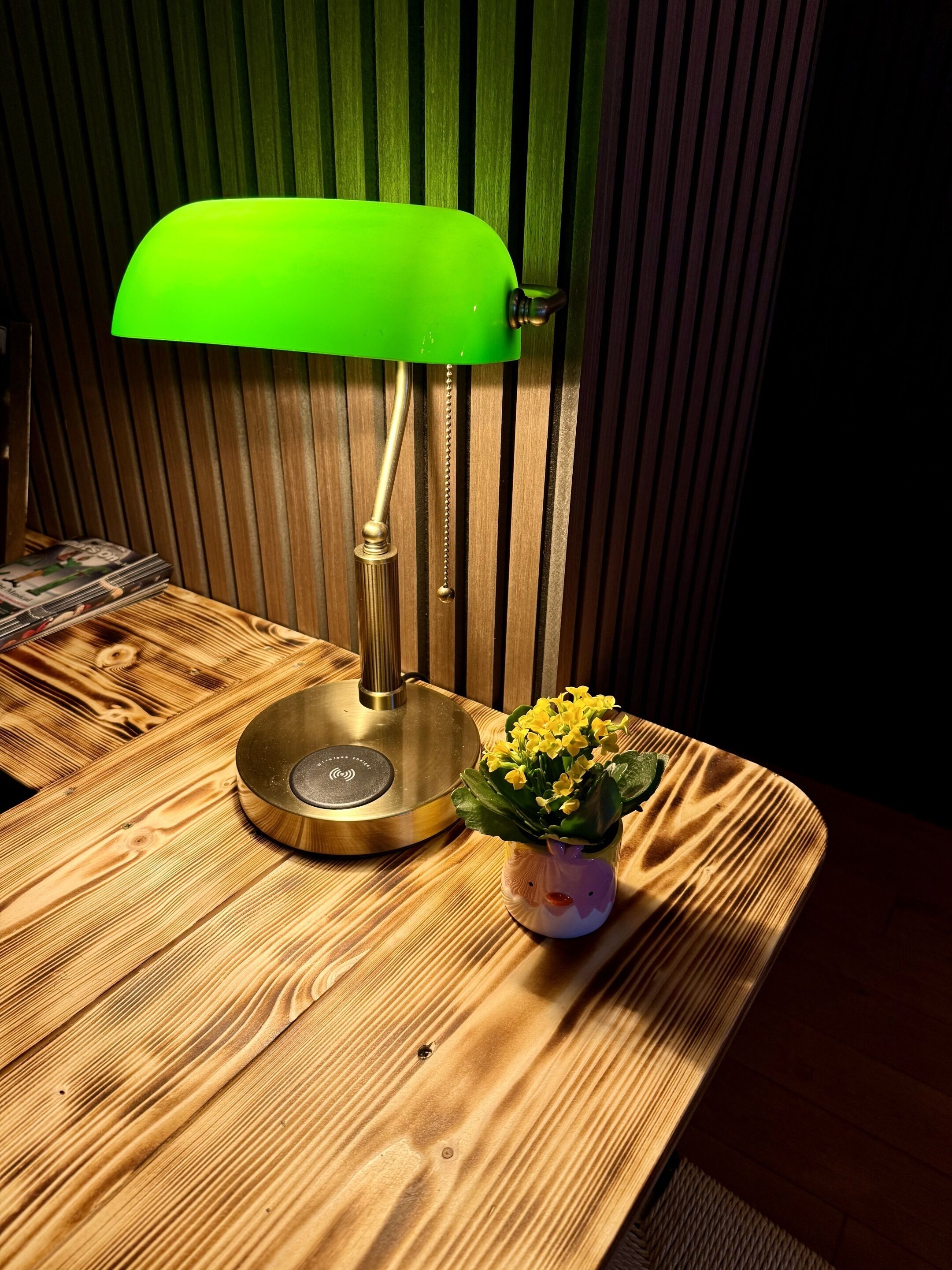A wooden table with a green desk lamp, a small vase with yellow flowers, and a stack of magazines.