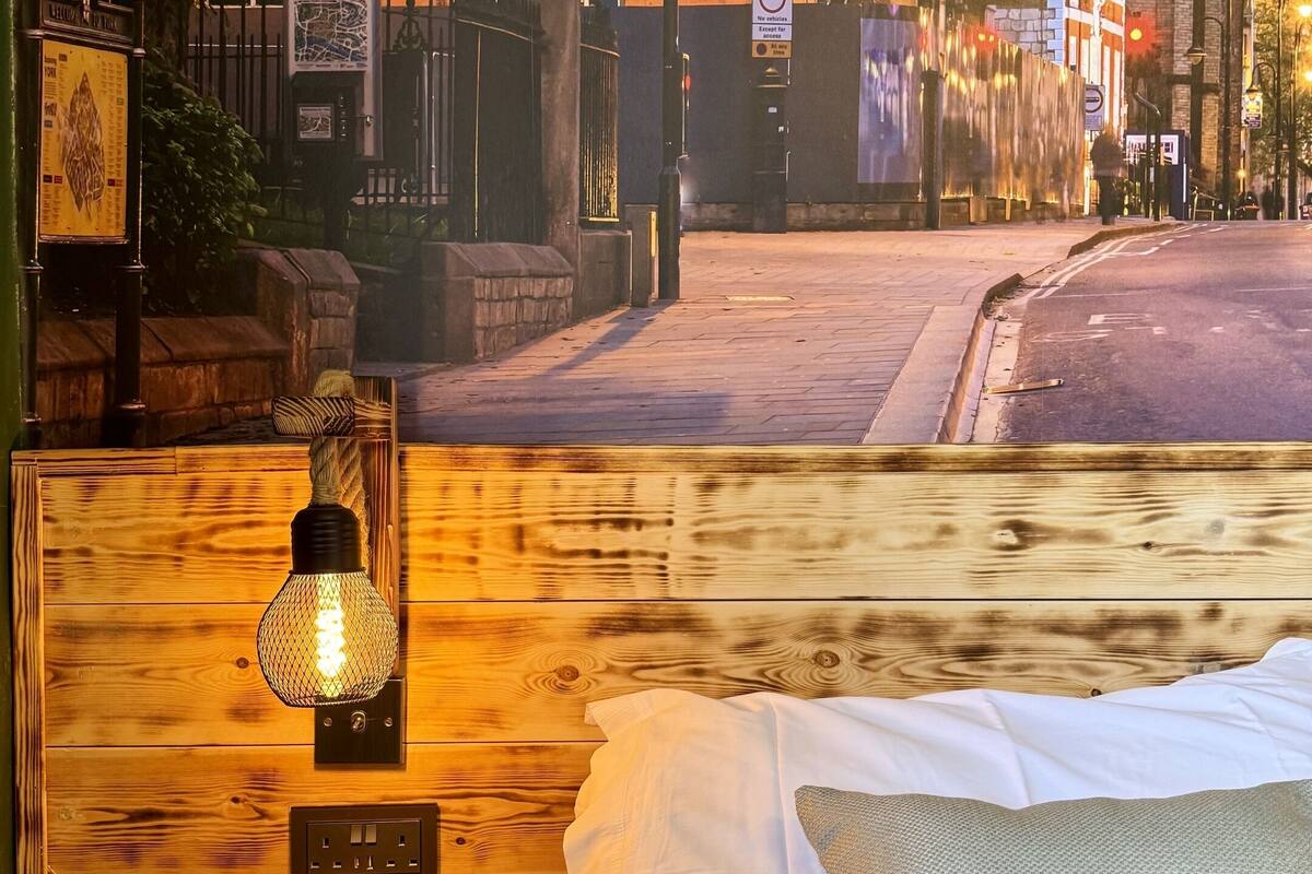 Una cama con sábanas blancas y un cojín texturizado, un cabecero de madera y una lámpara fijada a la pared.