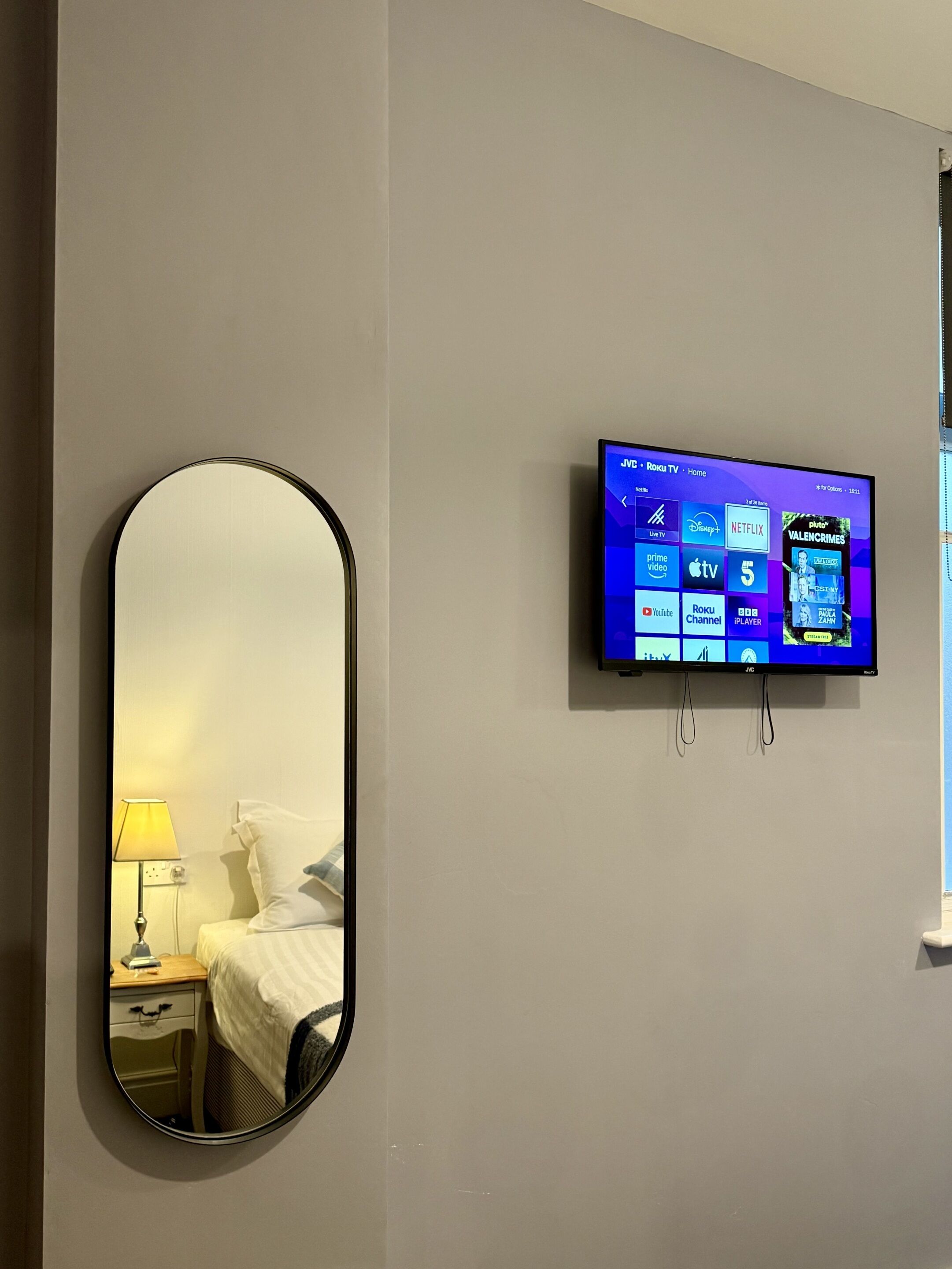 Habitación de hotel con cama, mesita de noche con lámpara, televisor montado en la pared mostrando servicios de streaming y un espejo grande en la pared.