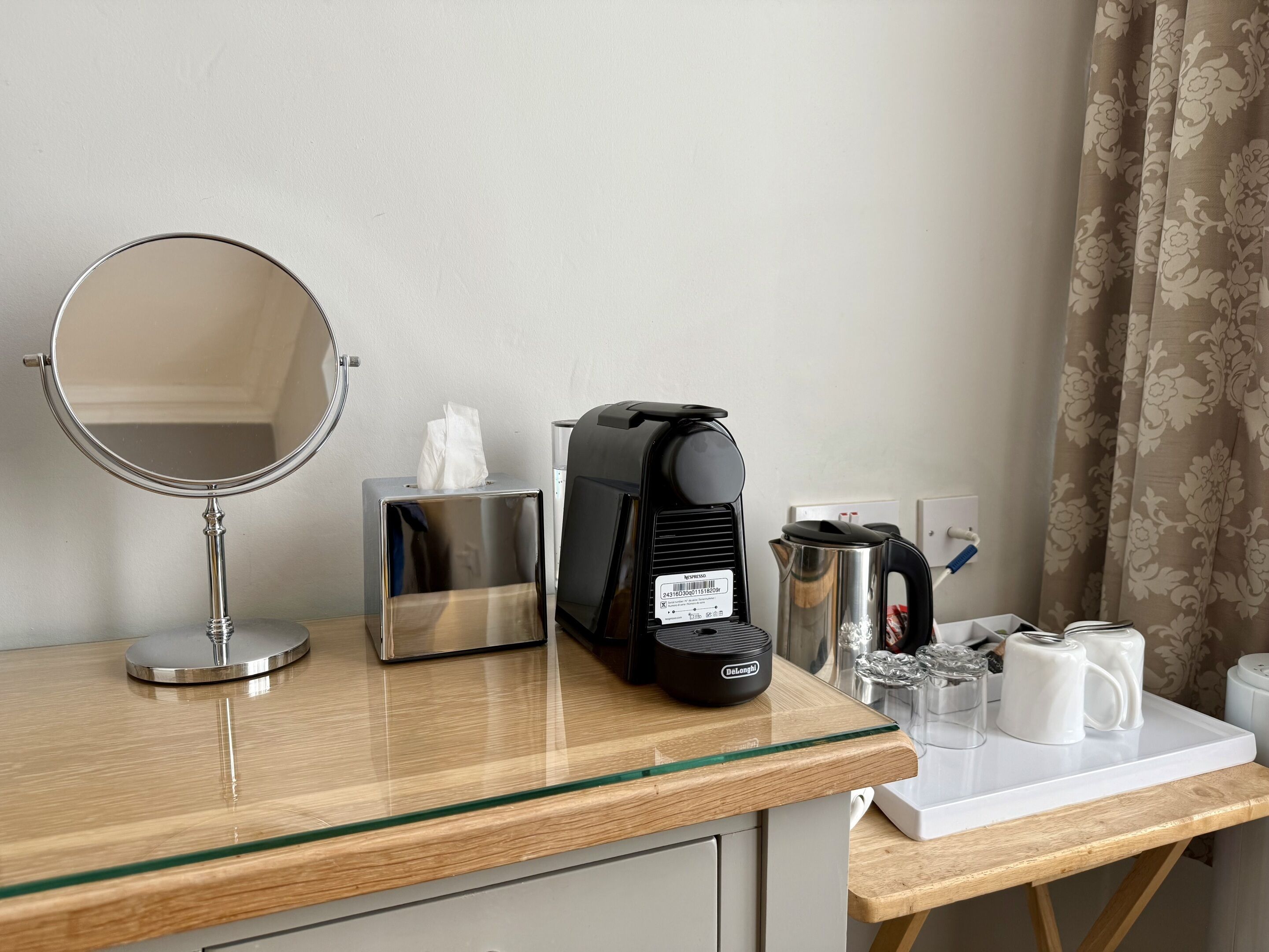A hotel room with a wooden vanity, a round mirror, a black De'Longhi coffee maker, a stainless steel tissue box, a white mug, and a kettle on a white countertop.