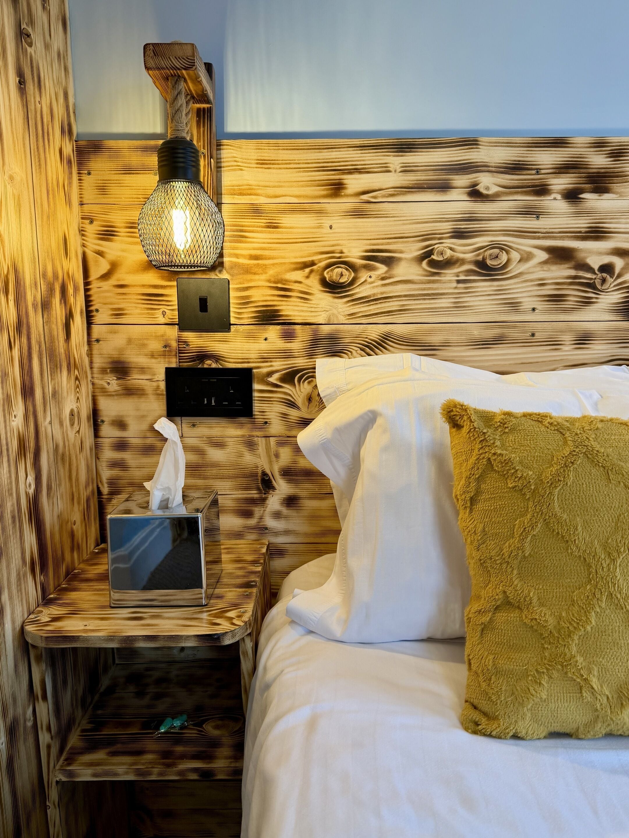 A wooden headboard with a textured finish, a bedside lamp, a tissue box, and a bed with white linens and a mustard-colored pillow.