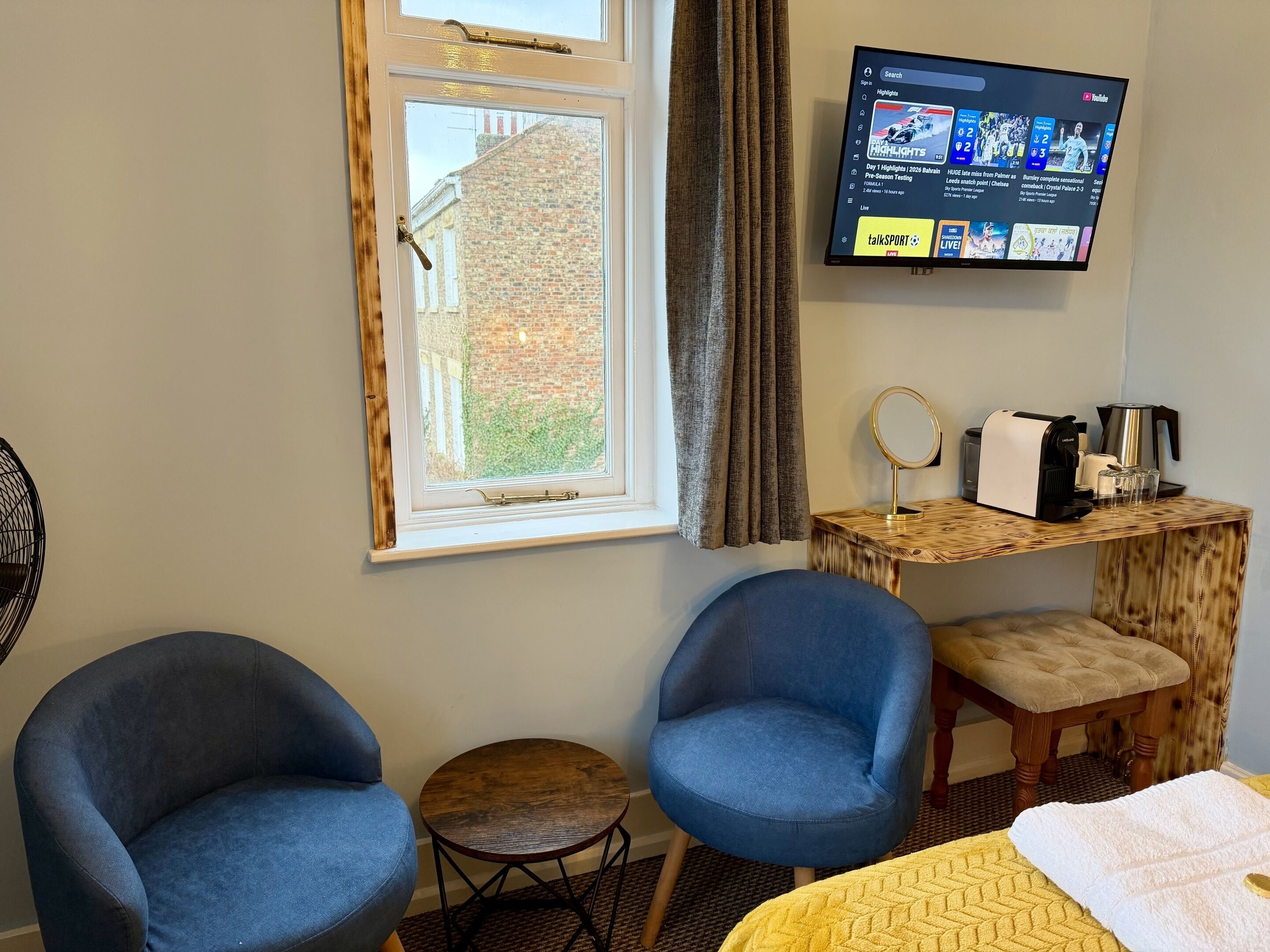 Habitación de hotel con dos sillones azules, una mesita redonda, una cafetera y un televisor mostrando momentos destacados de deportes.