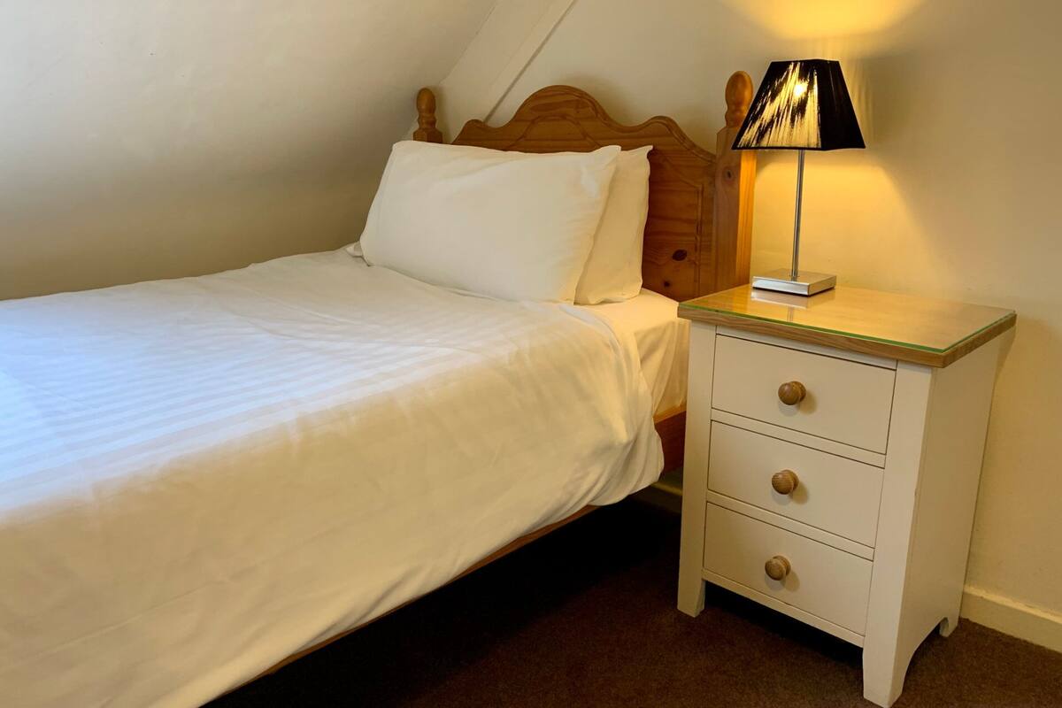 Una cama individual con ropa blanca, cabecera de madera, mesita de noche con cajones y una lámpara.