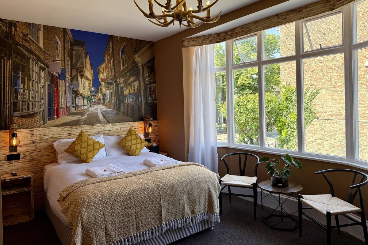 Habitación de hotel con un gran mural de una escena callejera, una cama con ropa de cama estampada, un candelabro y vistas a árboles a través de amplios ventanales.