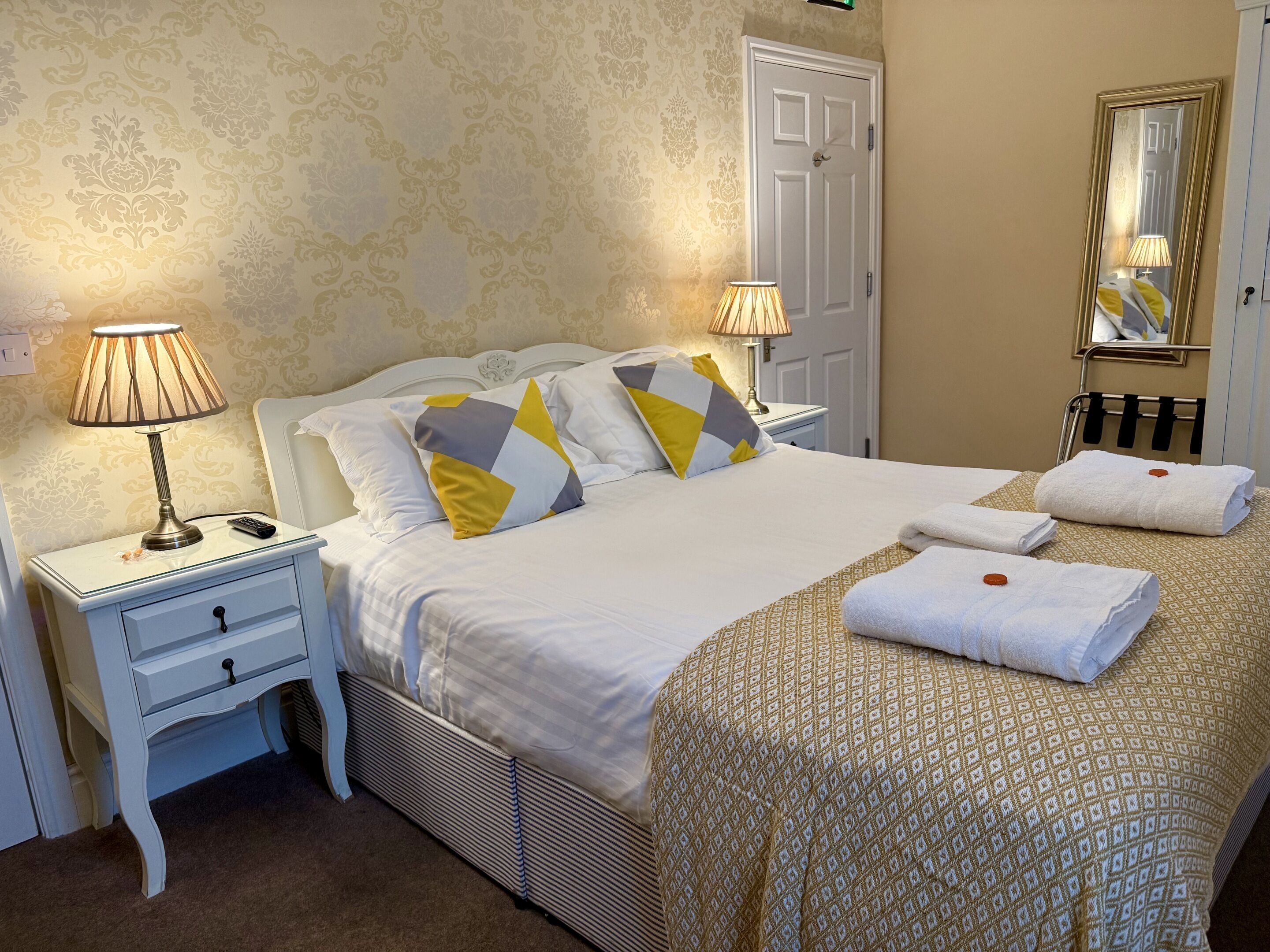 A neatly made bed with white bedding and patterned pillows, a bedside lamp, a small bedside table with a lamp, and a mirror on the wall.