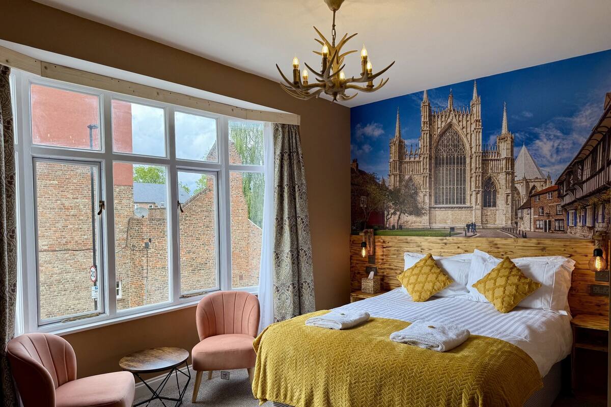 Un dormitorio con una cama grande, una silla rosa, una mesita y un mural de una catedral.
