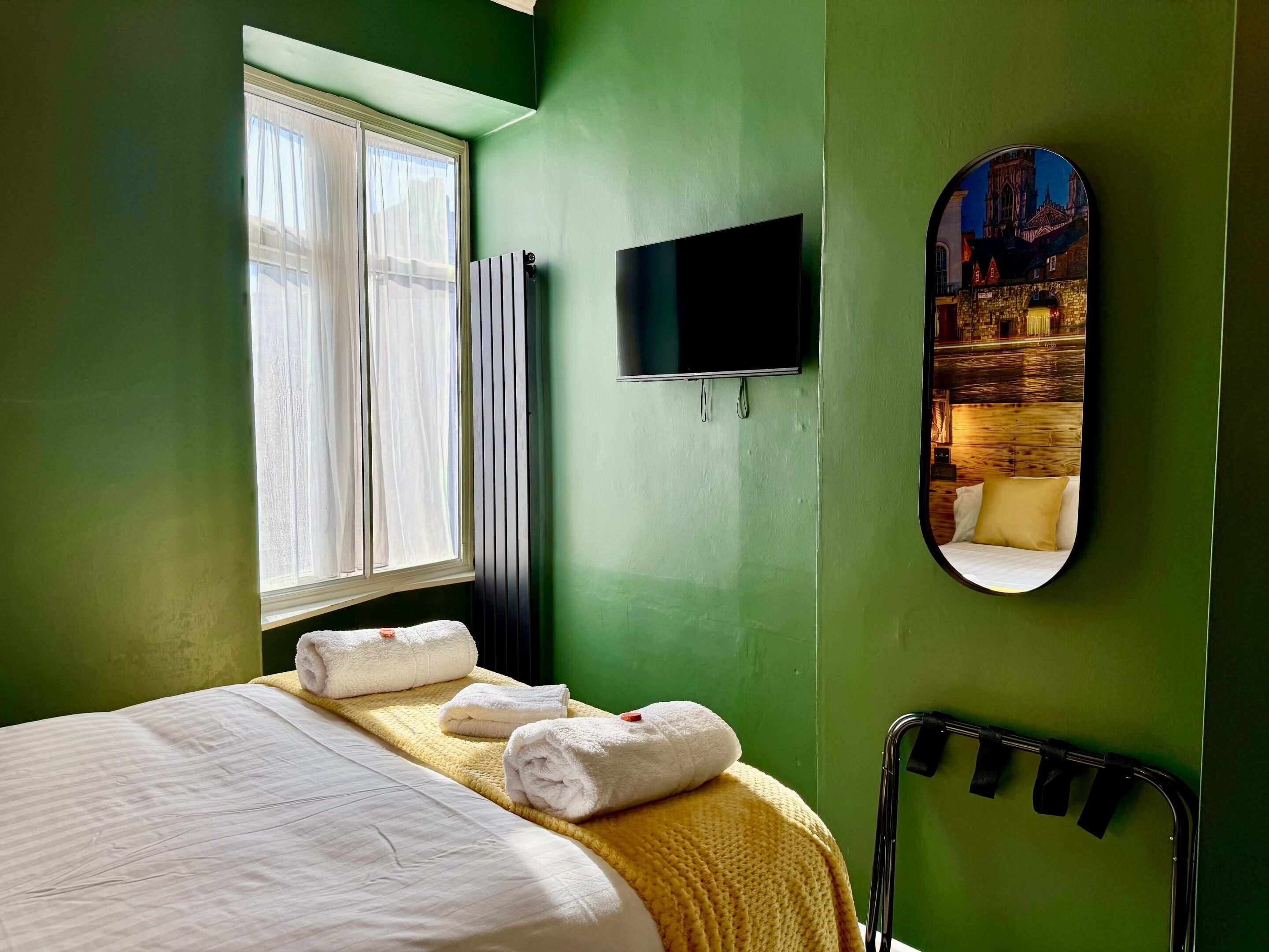 Ein Hotelzimmer mit Bett, einem Fenster mit Vorhängen, einem Flachbildfernseher und einem Spiegel, der ein Gebäude widerspiegelt.