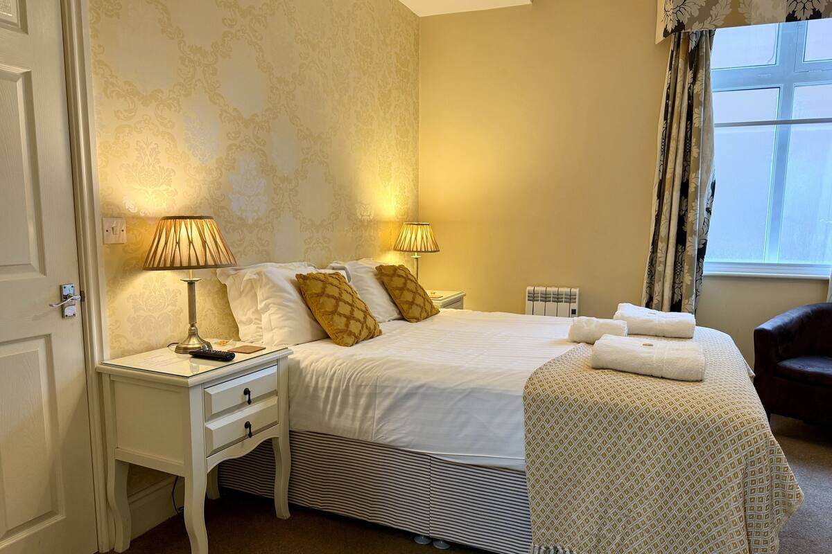 Un dormitorio ordenado con una cama, dos mesitas de noche con lámparas, un calefactor cerca de la cama y una ventana con cortinas.