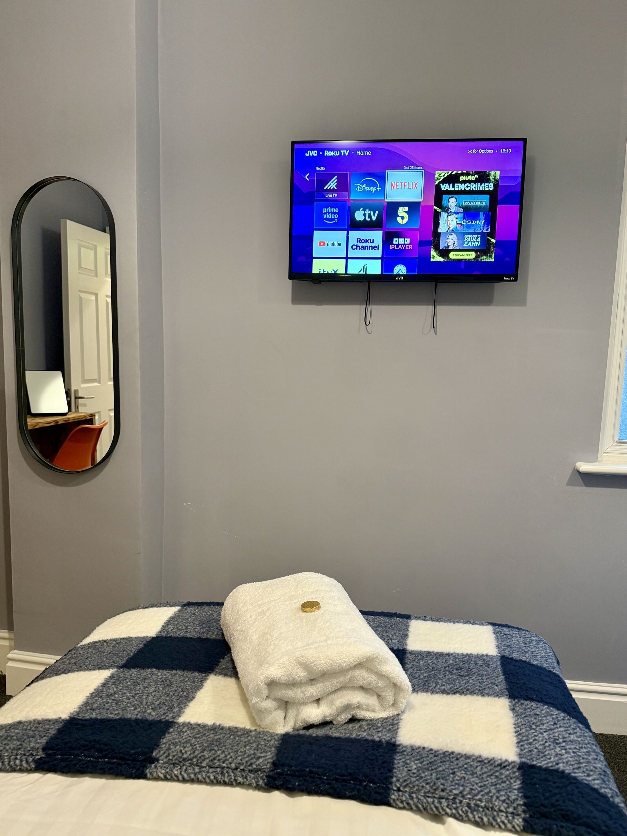 A bedroom with a bed, a TV mounted on the wall displaying various streaming services, a mirror, and a window.
