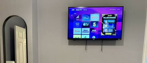 A bedroom with a bed, a TV mounted on the wall displaying various streaming services, a mirror, and a window.