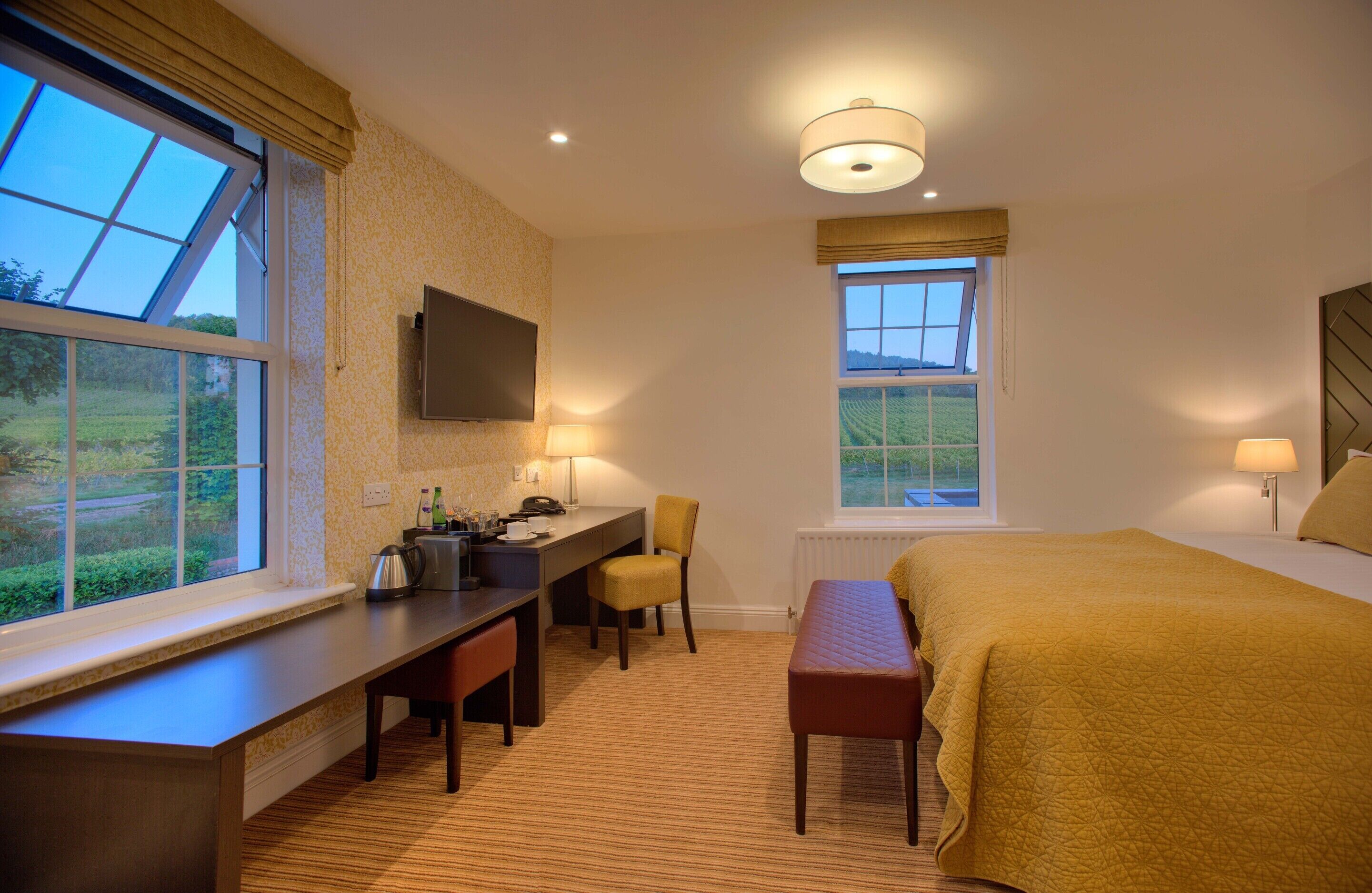 Una habitación de hotel con una cama grande, un escritorio con silla, un televisor en la pared y dos ventanas que ofrecen vistas al exterior.
