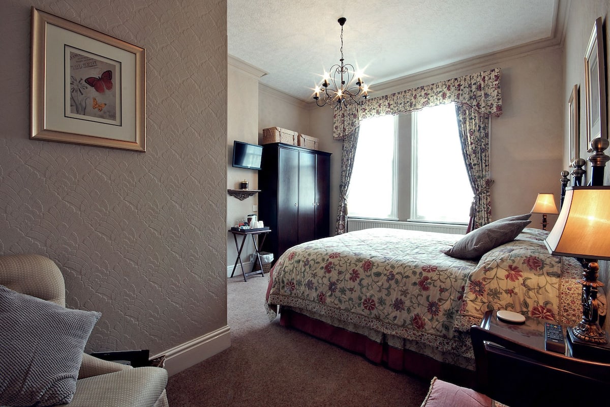 A bedroom with a bed, a chair, a dresser, a television, and a window with curtains.