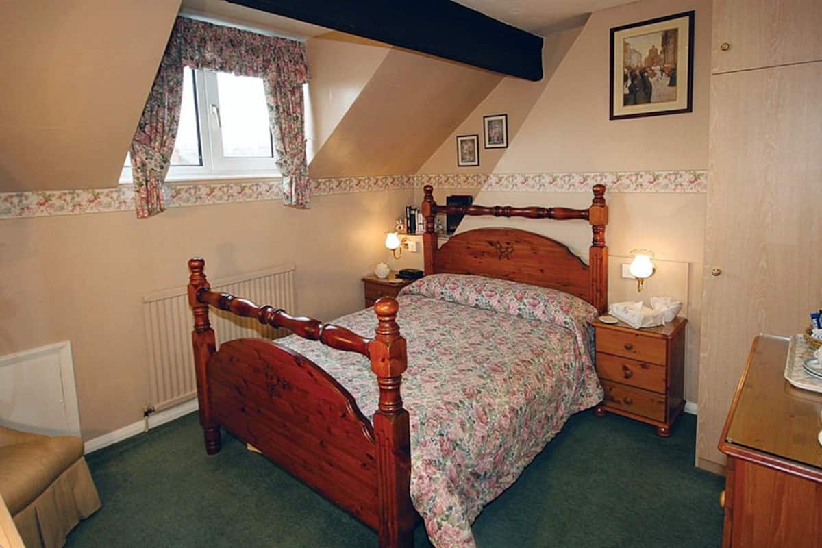 A bedroom with a wooden four-poster bed, two bedside tables with lamps, a dresser, and a window with floral curtains.
