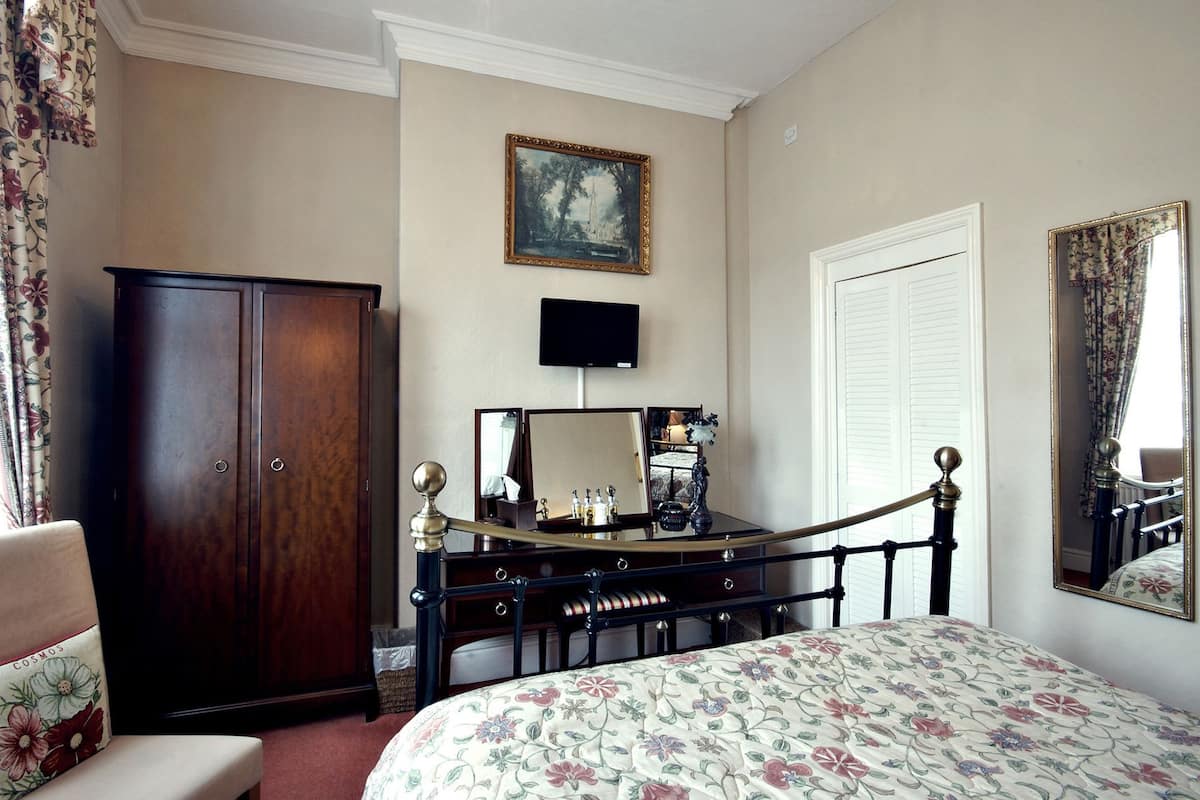 A hotel room with a bed, a wardrobe, a vanity with a mirror, a television, and a painting on the wall.