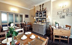 Ein Essbereich mit einem Holztisch, gedeckt für vier Personen, umgeben von Stühlen. Ein Steinkamin mit Sims befindet sich über dem Tisch, und ein Schrank mit diversen Gegenständen darauf.