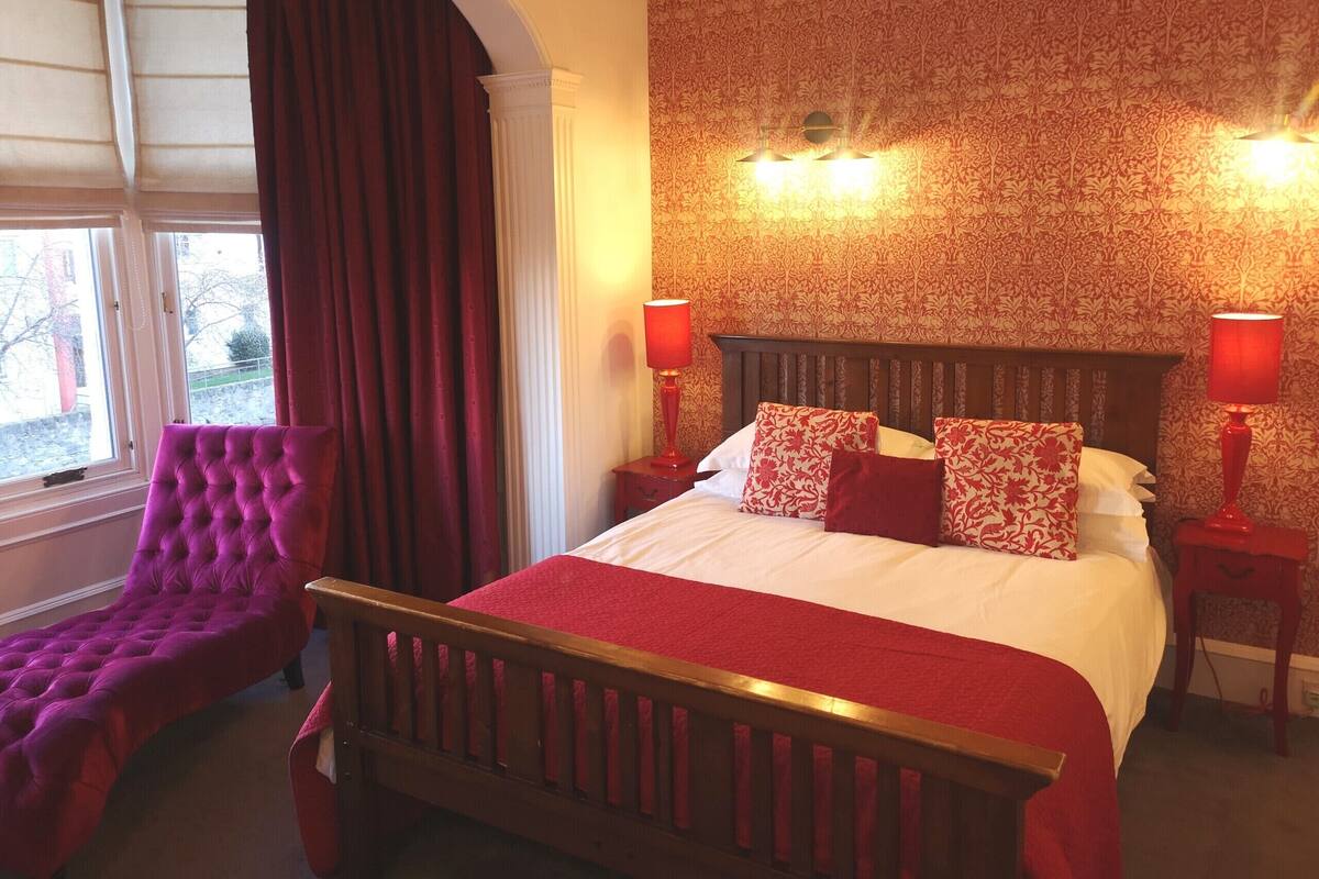 Une chambre d’hôtel avec un grand lit, deux tables de chevet, un fauteuil rouge recouvert de tissu et une fenêtre avec des rideaux.