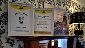 A hotel reception area with framed awards for breakfast and guest accommodation, a vase of tulips, and a red chair. - Fraoch House (Edinburgh)