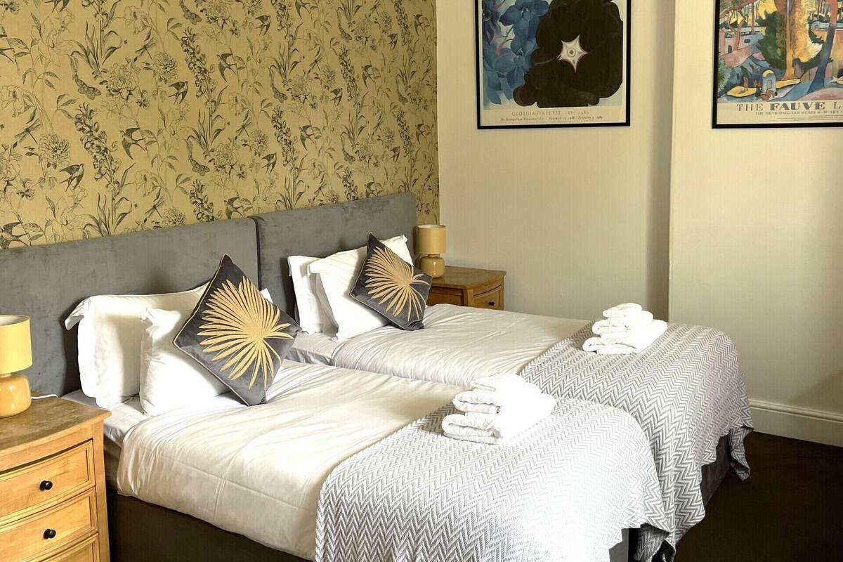 Un lit double avec une tête de lit grise, deux tables de chevet, une commode, un lustre et un papier peint à motifs floraux.