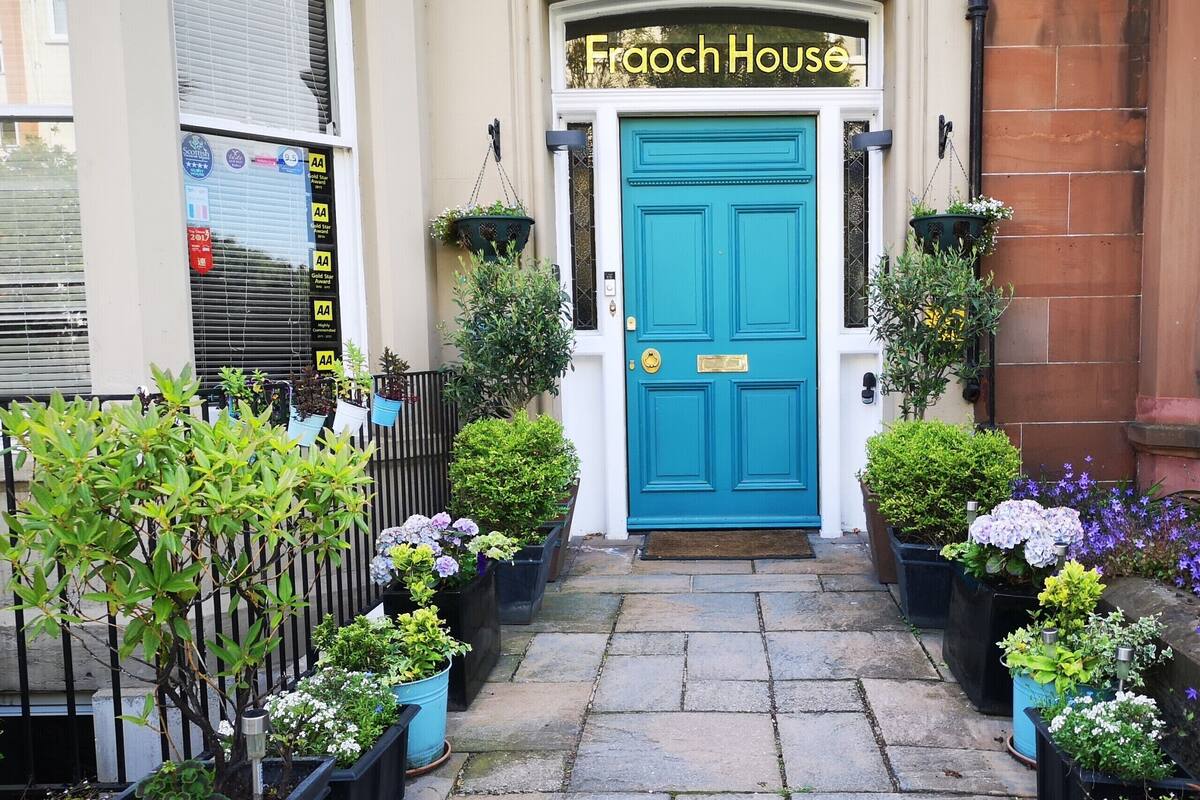 L’entrée d’un immeuble, avec une porte bleue portant l’inscription « Frooch House », et des plantes en pot de chaque côté.