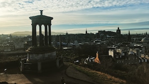 A historical monument with columns, surrounded by a cityscape with various buildings and a clear sky. - Fraoch House (Edinburgh)