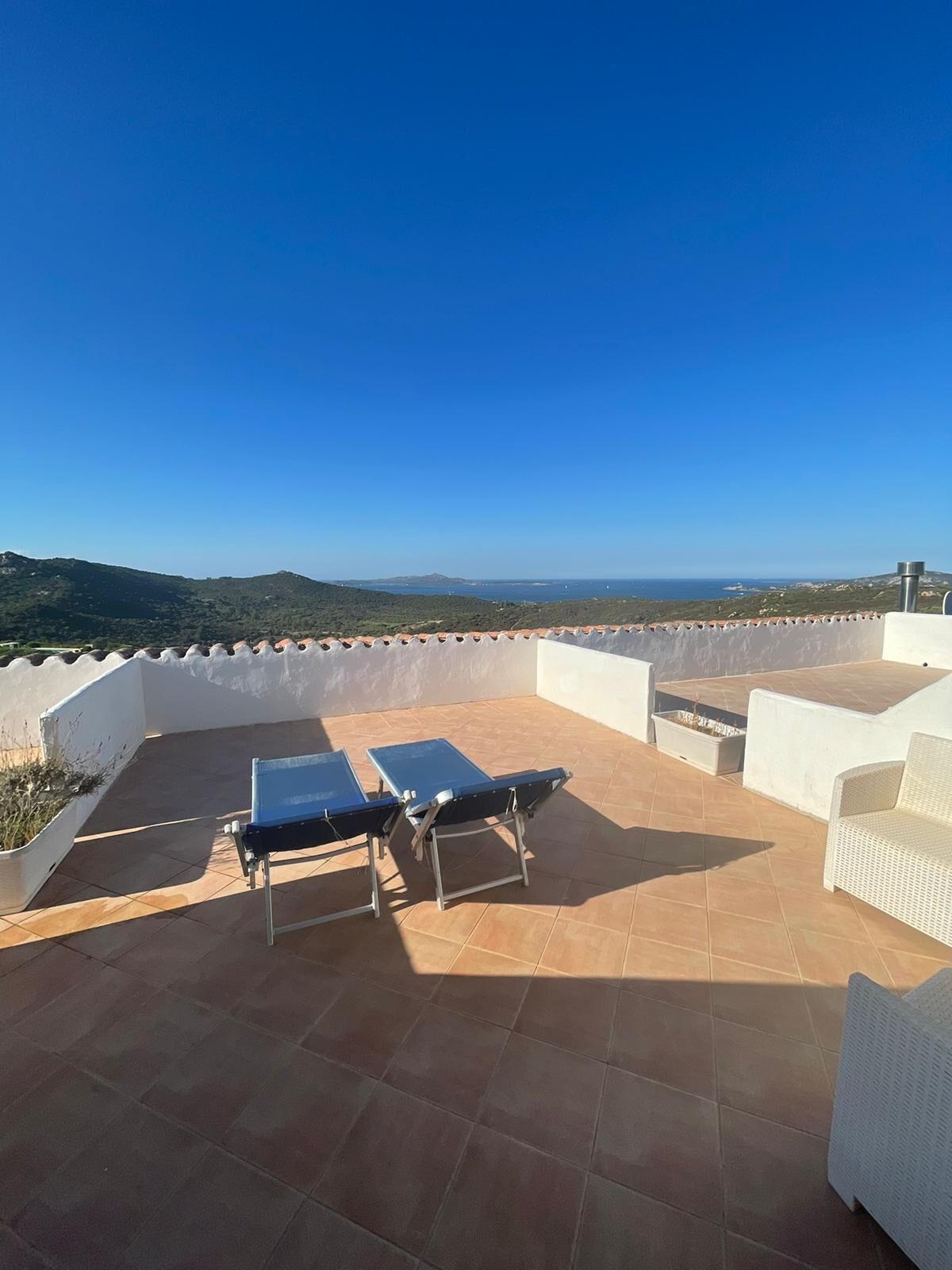 A rooftop terrace with two blue sun loungers, a white wicker sofa, and a view of the sea and hills.