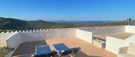 A rooftop terrace with two blue sun loungers, a white wicker sofa, and a view of the sea and hills.