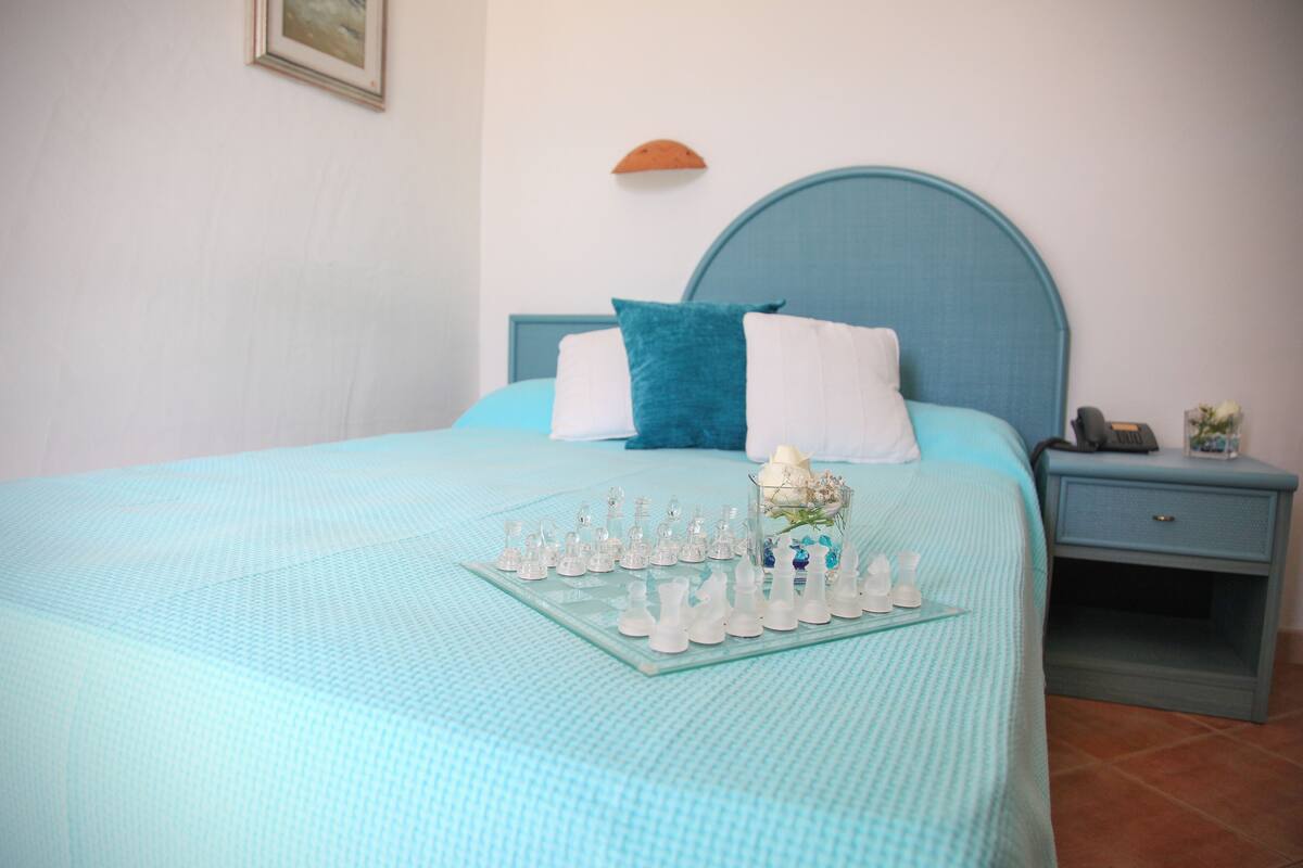 A neatly made bed with a blue headboard, a glass chessboard, and a nightstand with a lamp and a vase of flowers.