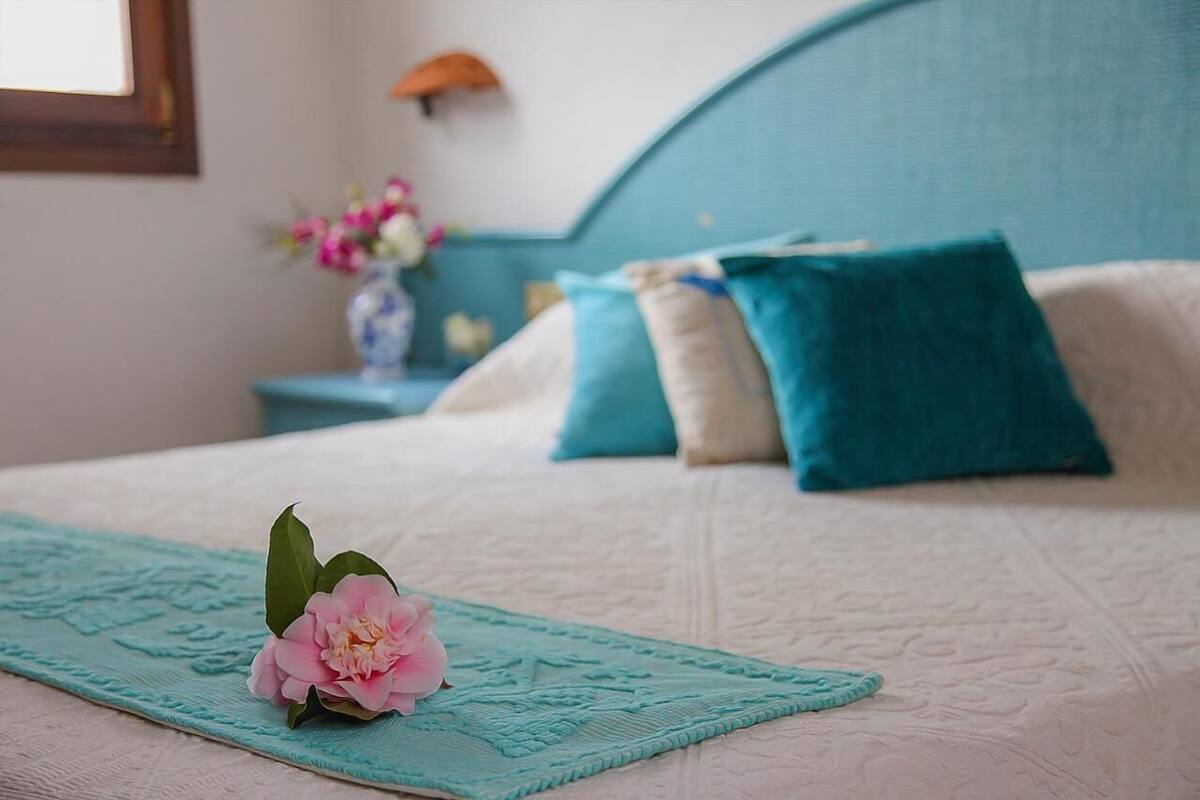 A neatly made bed with a teal bed runner, a single pink flower, and two pillows in teal and white.