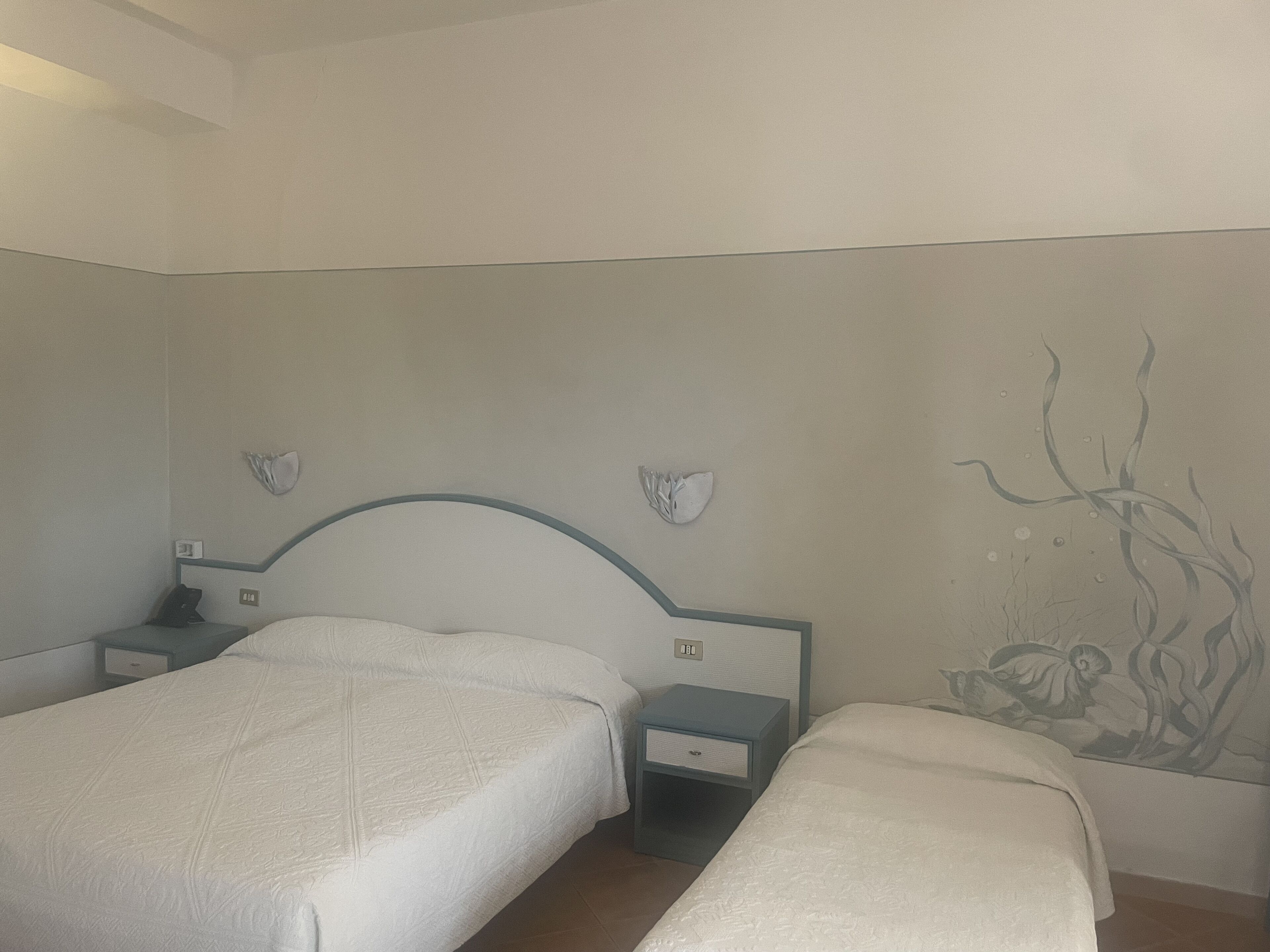 A hotel room with two single beds, a nightstand with a lamp, and a wall mural.
