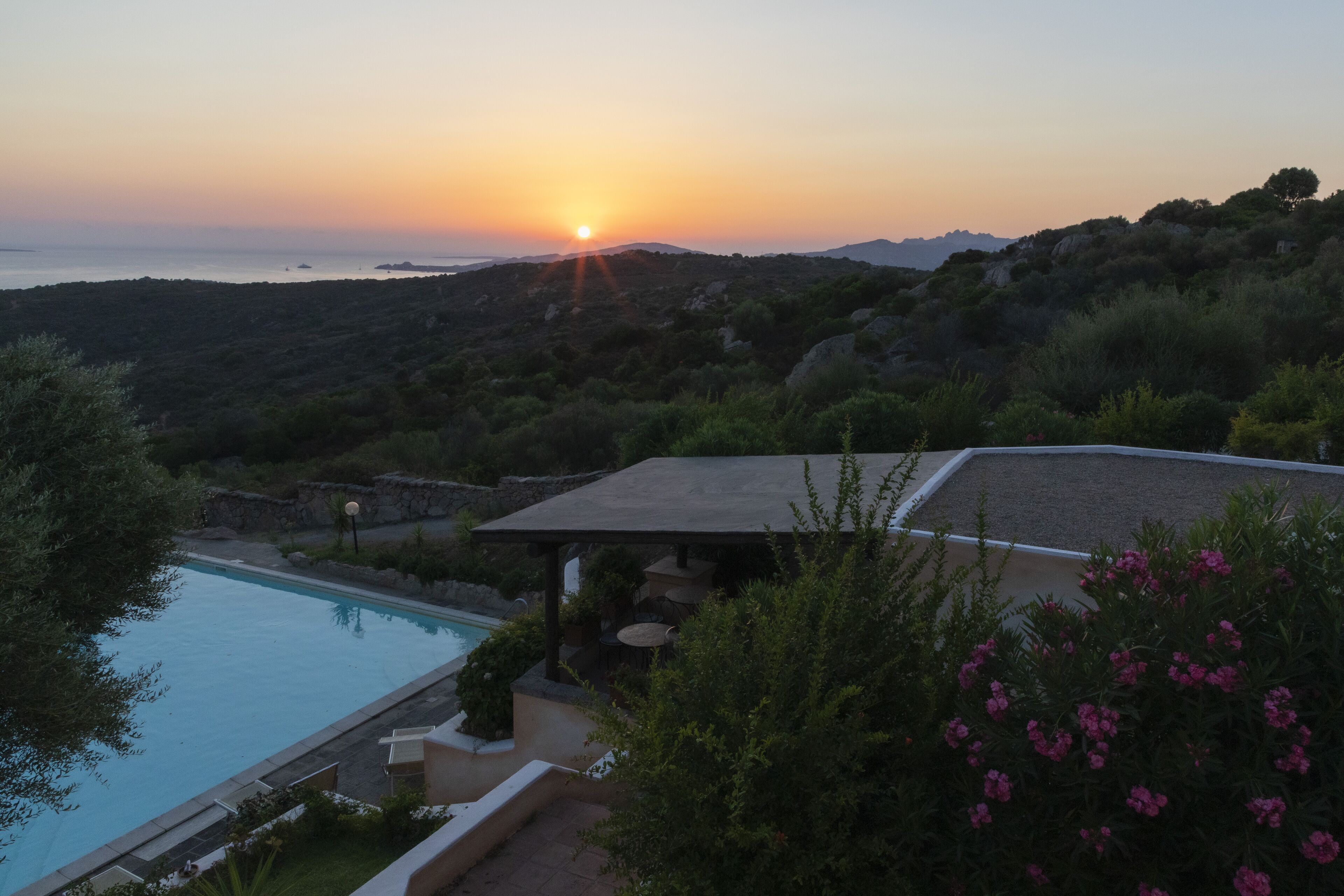 A pool area with a view of the sunset over the sea, surrounded by greenery and hills.