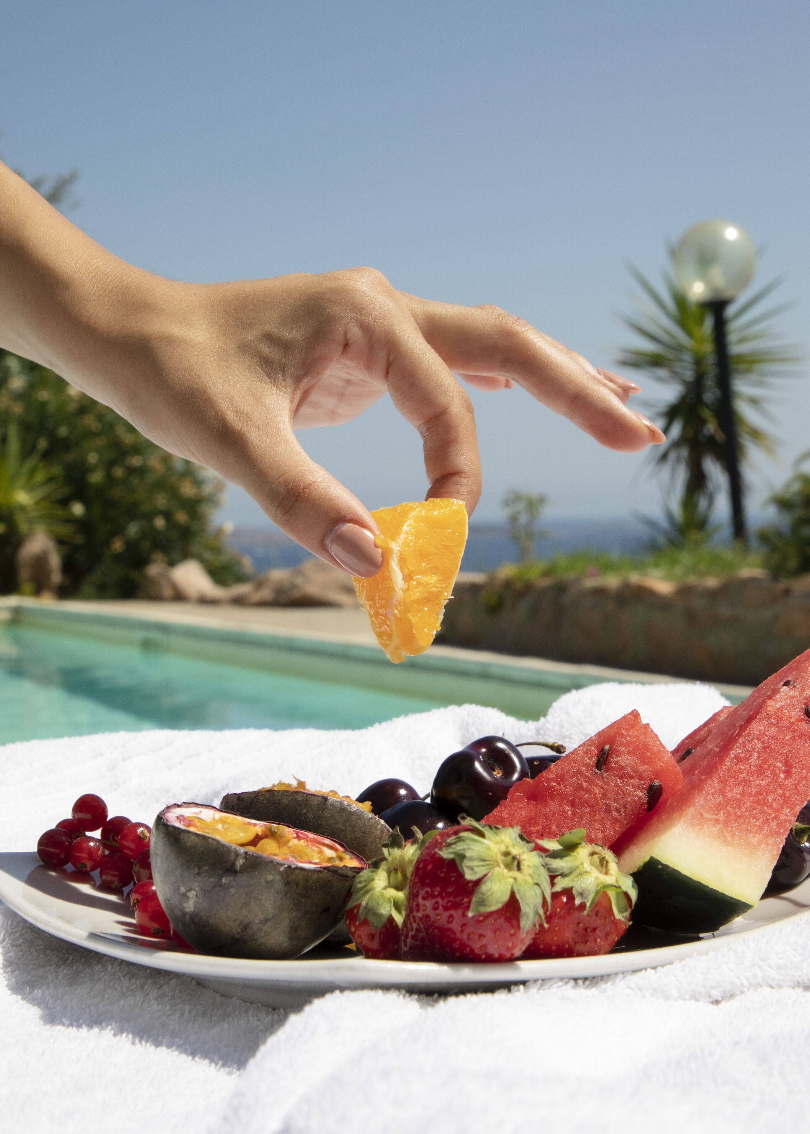 Una mano sostiene un gajo de naranja sobre un plato de frutas, que incluye sandía, fresas y uvas, con una piscina y palmeras al fondo.