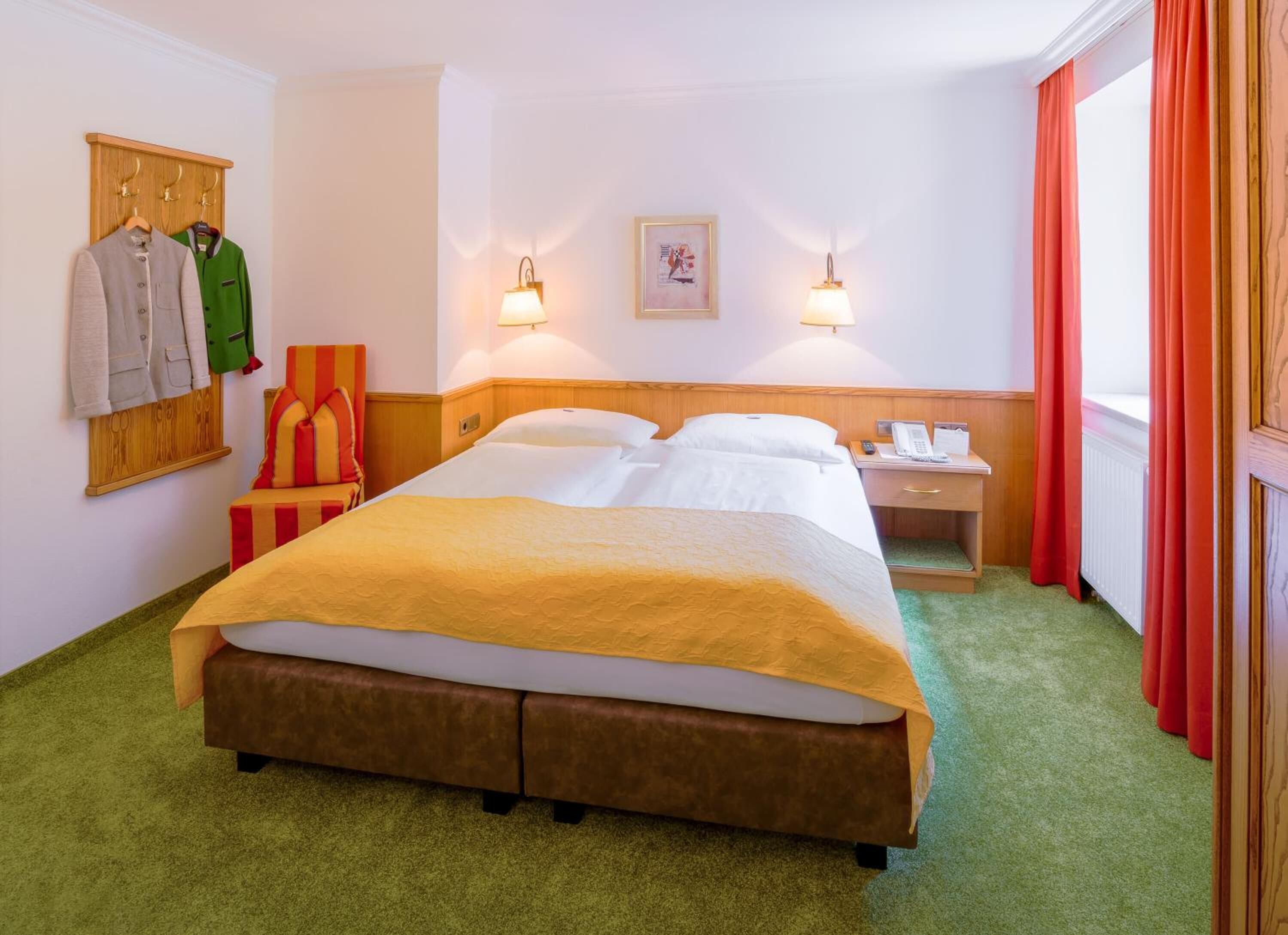 A hotel room with a bed, a nightstand, a lamp, a telephone, and a coat rack with jackets.