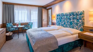 A hotel room with a large bed, a desk with a chair, a window with curtains, and a headboard with a floral pattern. - Hotel Jakober (Tux)