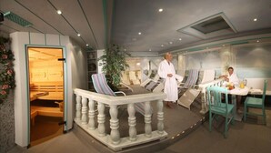 A spa room with a massage chair, a small table with a drink, and a person in a robe. - Hotel Jakober (Tux)
