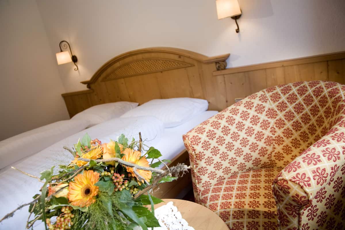 Habitación de hotel con cabecera de madera, dos camas, un sillón estampado y un arreglo floral en la mesita de noche.