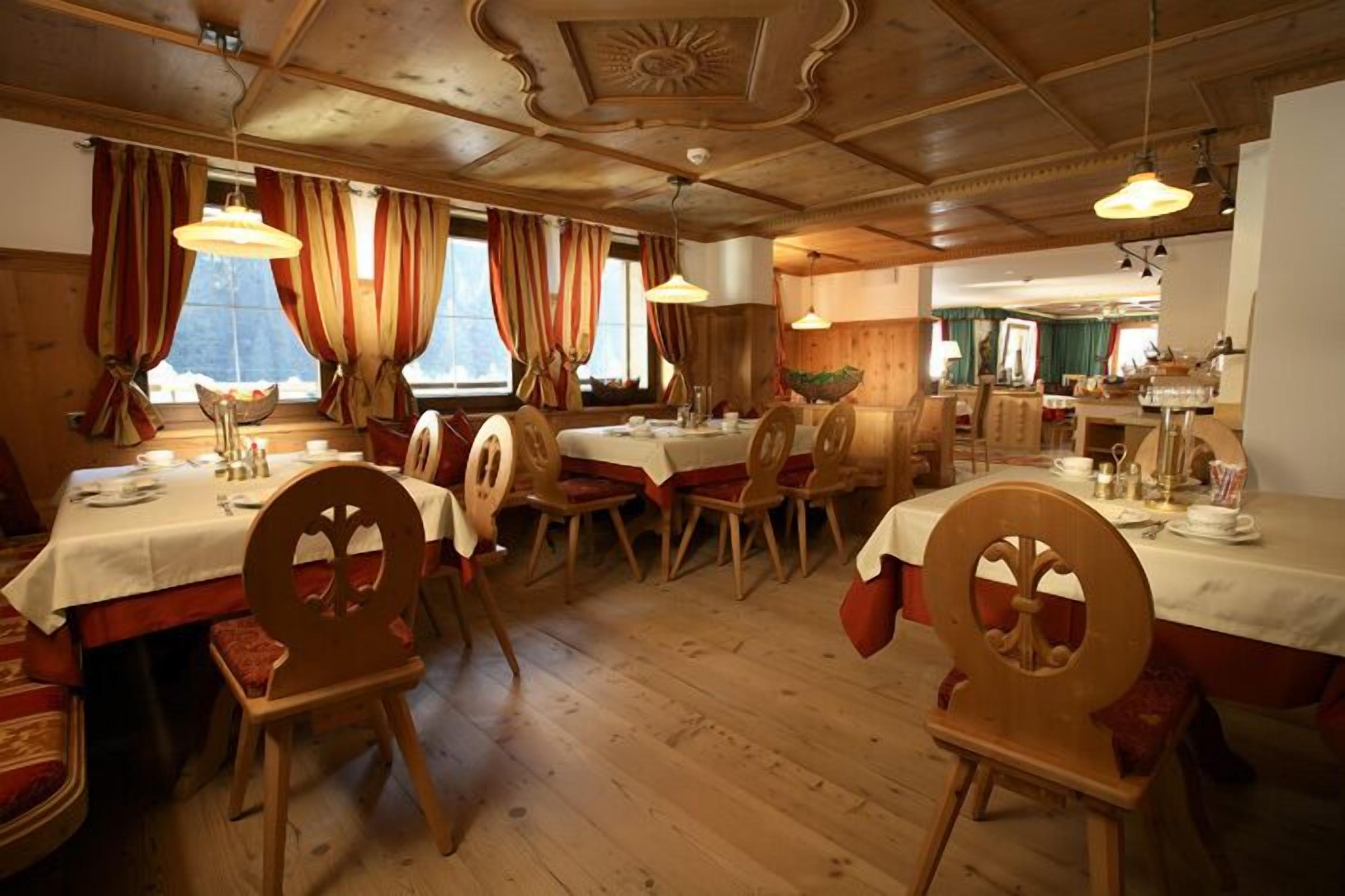 Uma sala de jantar com mesas e cadeiras de madeira, janelas amplas com cortinas e teto de madeira.