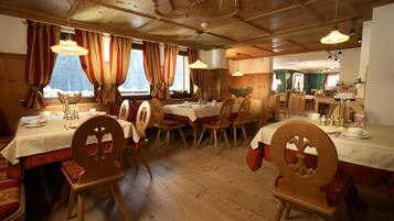 Uma sala de jantar com mesas e cadeiras de madeira, janelas amplas com cortinas e teto de madeira.