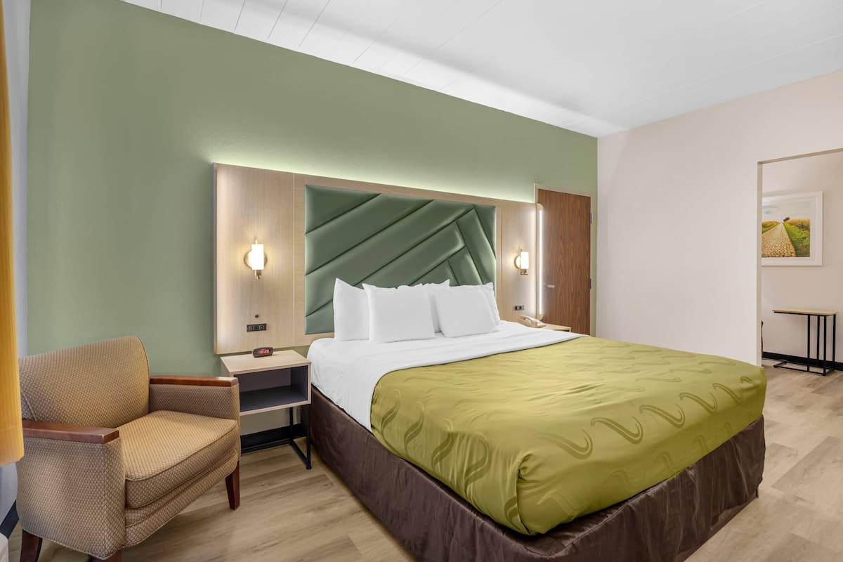A hotel room with a bed, a chair, a nightstand, and a wall-mounted lamp.