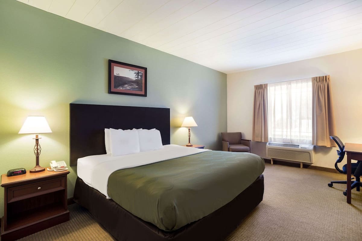 A hotel room with a bed, bedside lamps, a chair, a desk, a window with curtains, and a framed picture on the wall.