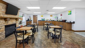 A restaurant with a stone fireplace, tiled floor, wooden tables and chairs, a kitchen area, and a flat-screen TV mounted on the wall. - Quality Inn & Suites Monterey I-40 (Monterey)