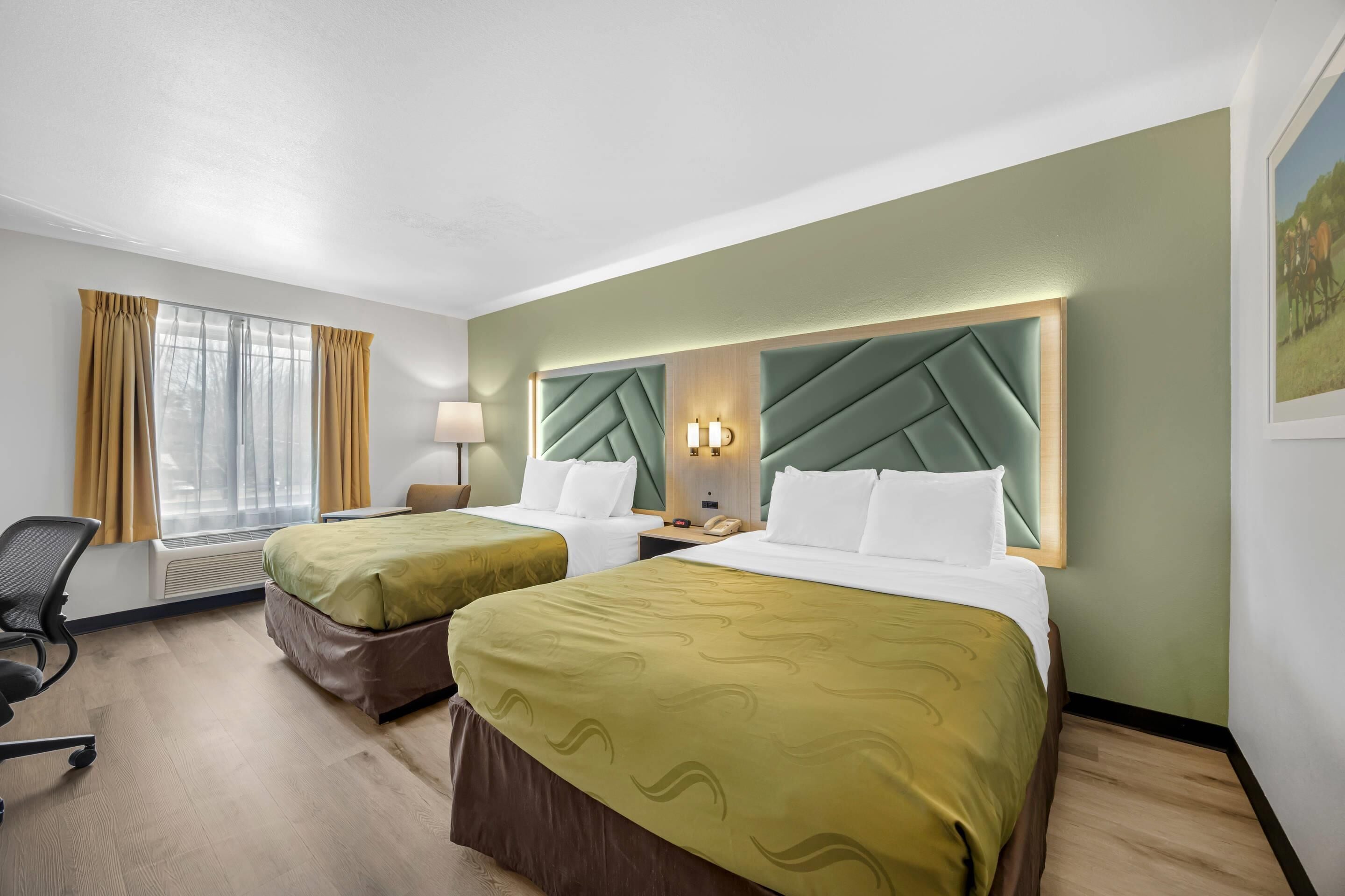 A hotel room with two beds, a desk, a chair, a window with curtains, and a wall-mounted lamp.