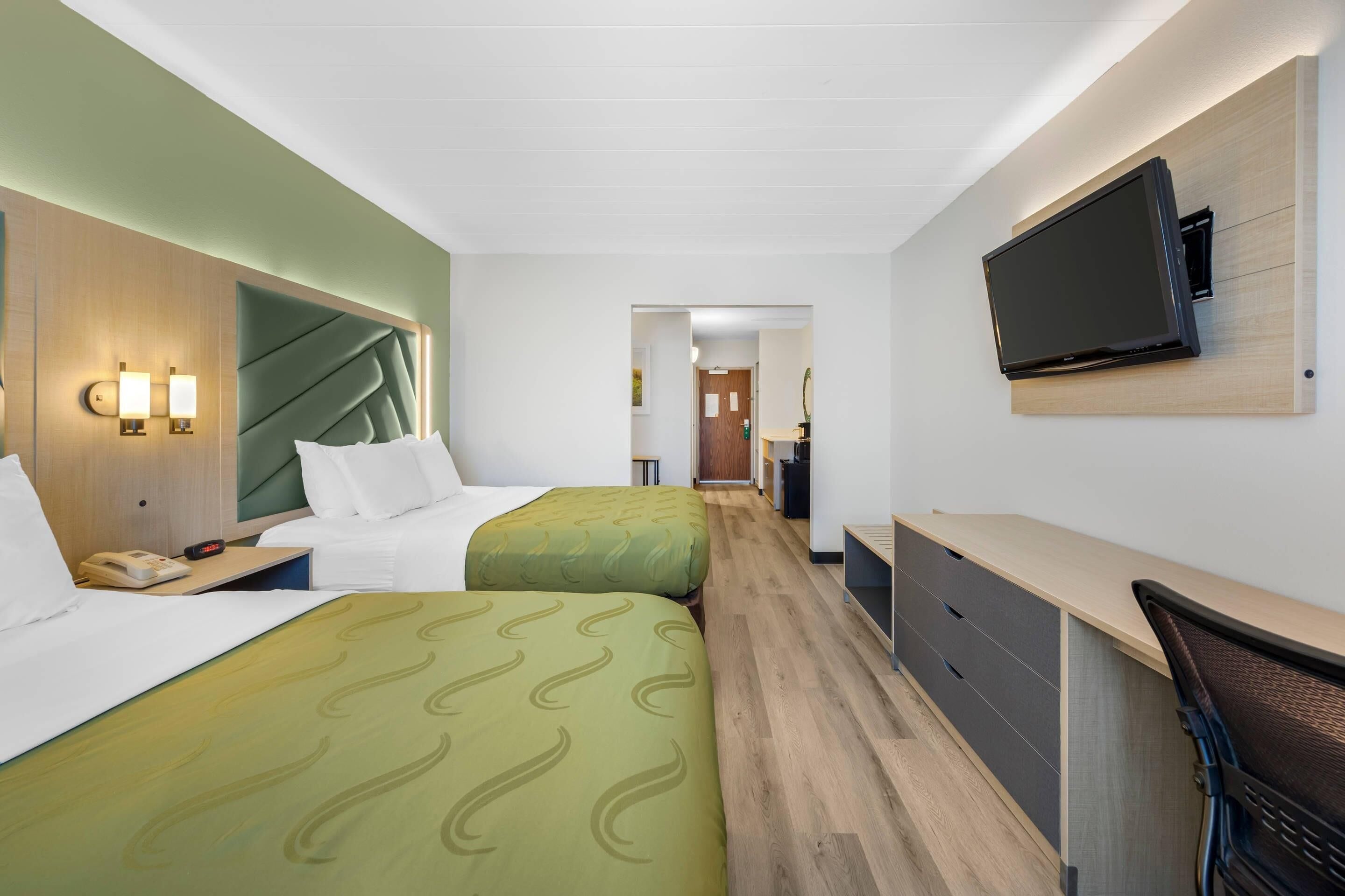 Habitación de hotel con dos camas, un escritorio, una silla, un televisor montado en la pared y piso de madera.