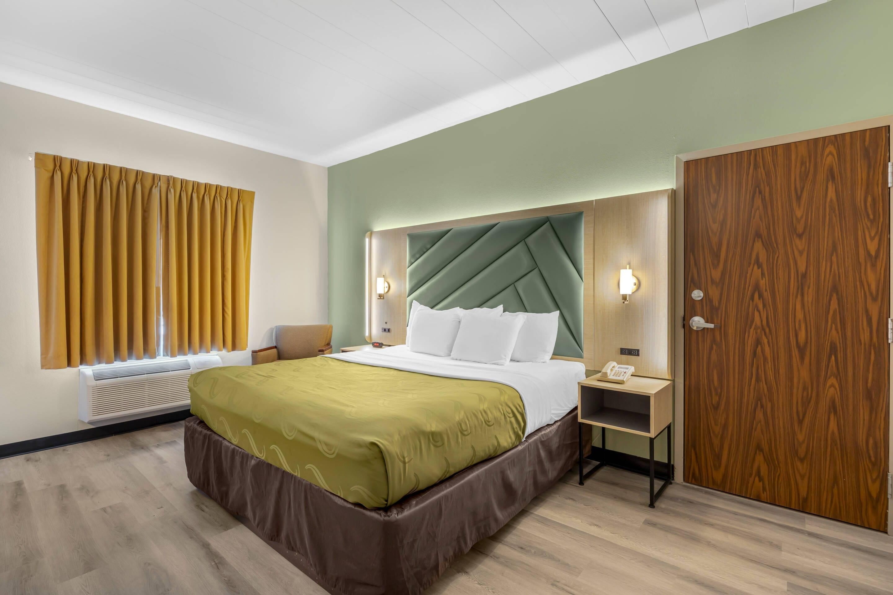 Una habitación de hotel con cama, mesita de noche, armario de madera y ventana con cortinas.