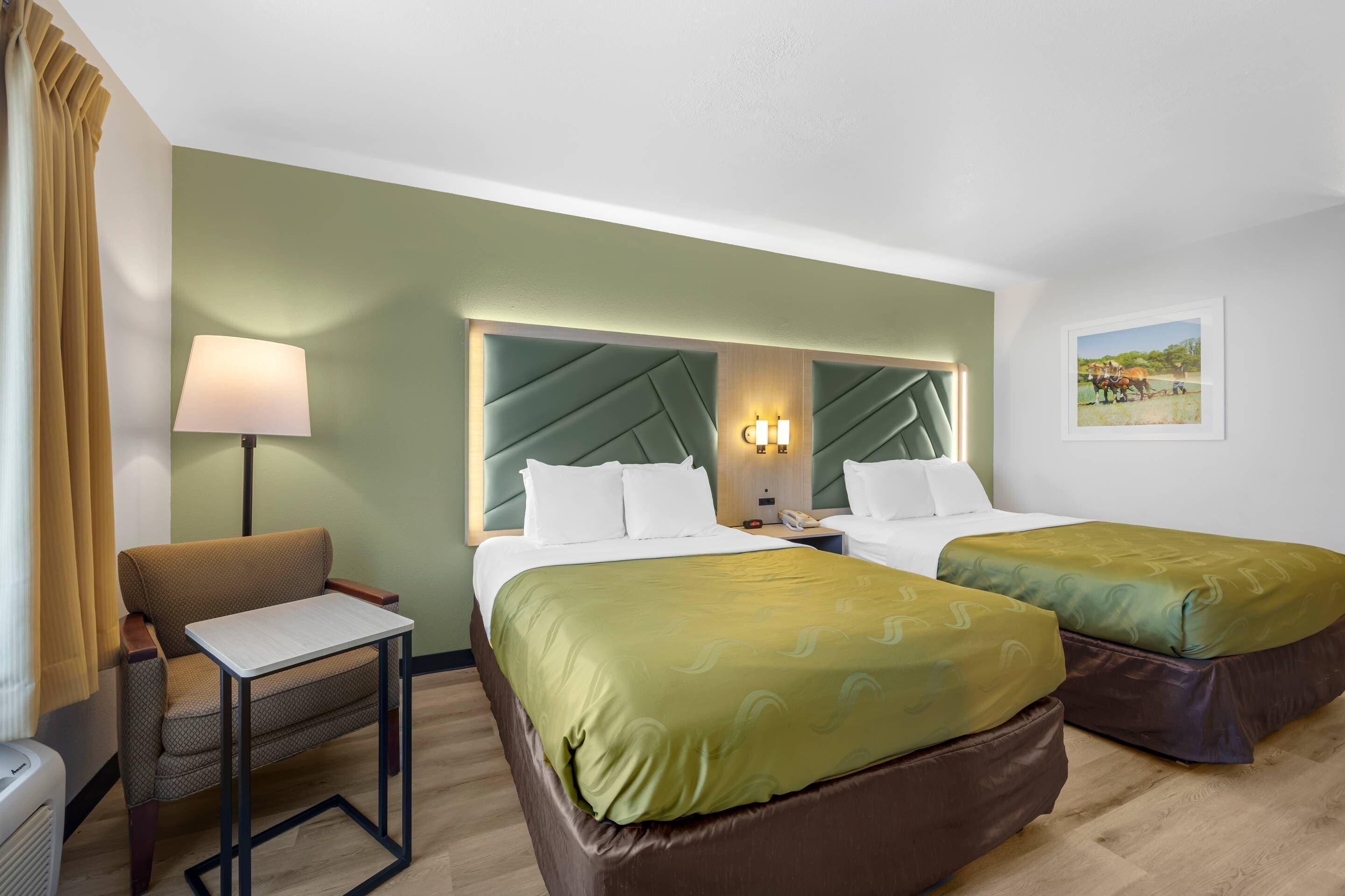 A hotel room with two beds, a desk, a chair, a lamp, and a painting on the wall.