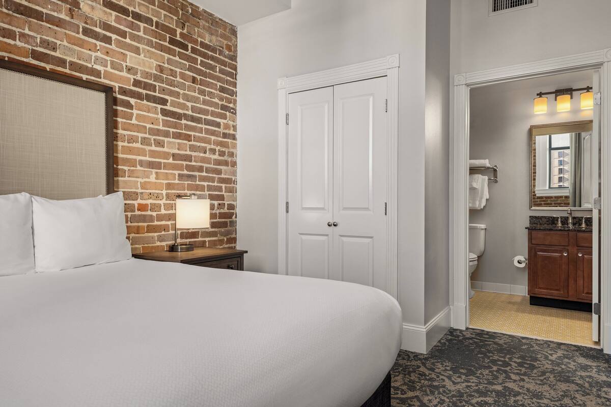 Une chambre d’hôtel avec un mur d’accent en briques, un grand lit avec du linge de lit blanc, une table de chevet avec une lampe, une porte blanche et une vue sur la salle de bain.