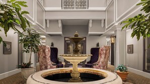 A grand hallway with a central fountain, flanked by purple armchairs and potted plants.